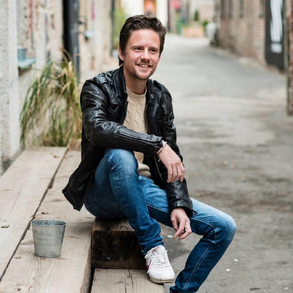 David sitting outdoors on a wooden bench, smiling casually in a leather jacket and jeans.